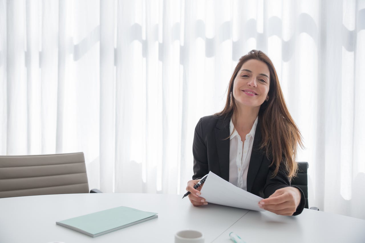 award-img Smiling businesswoman in a blazer holding paper at an office table indoors.